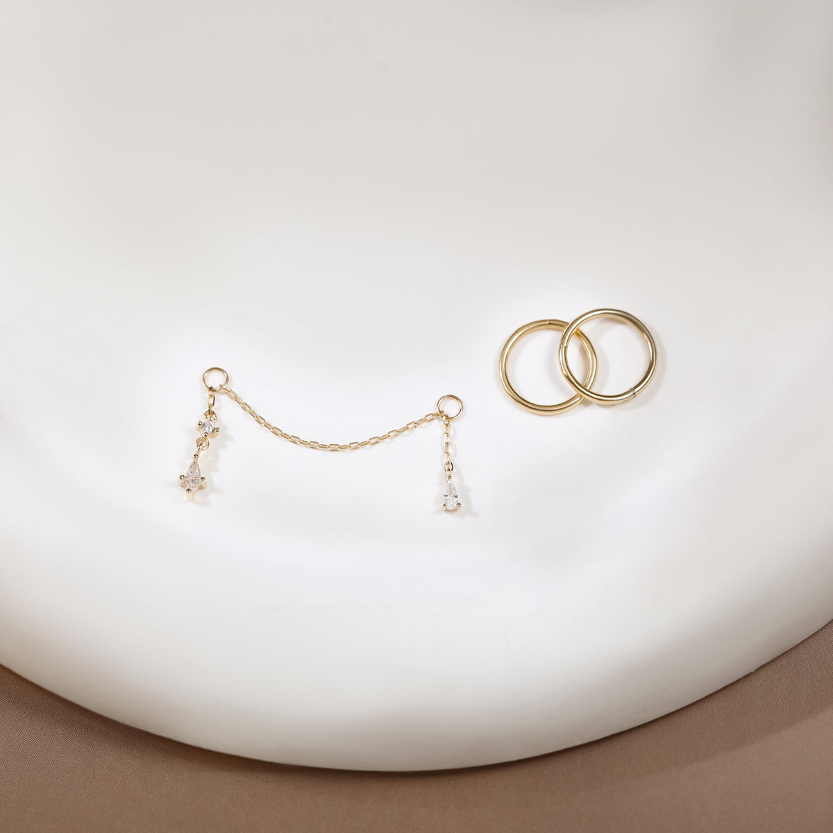 A pair of gold-toned earrings displayed on a white curved surface, including two interlocking hoop-style rings and a delicate chain bridge with small dangling clear crystal drops.