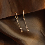 A pair of gold-toned dangle earrings with small chain drops and triangular charms, photographed on draped brown fabric.