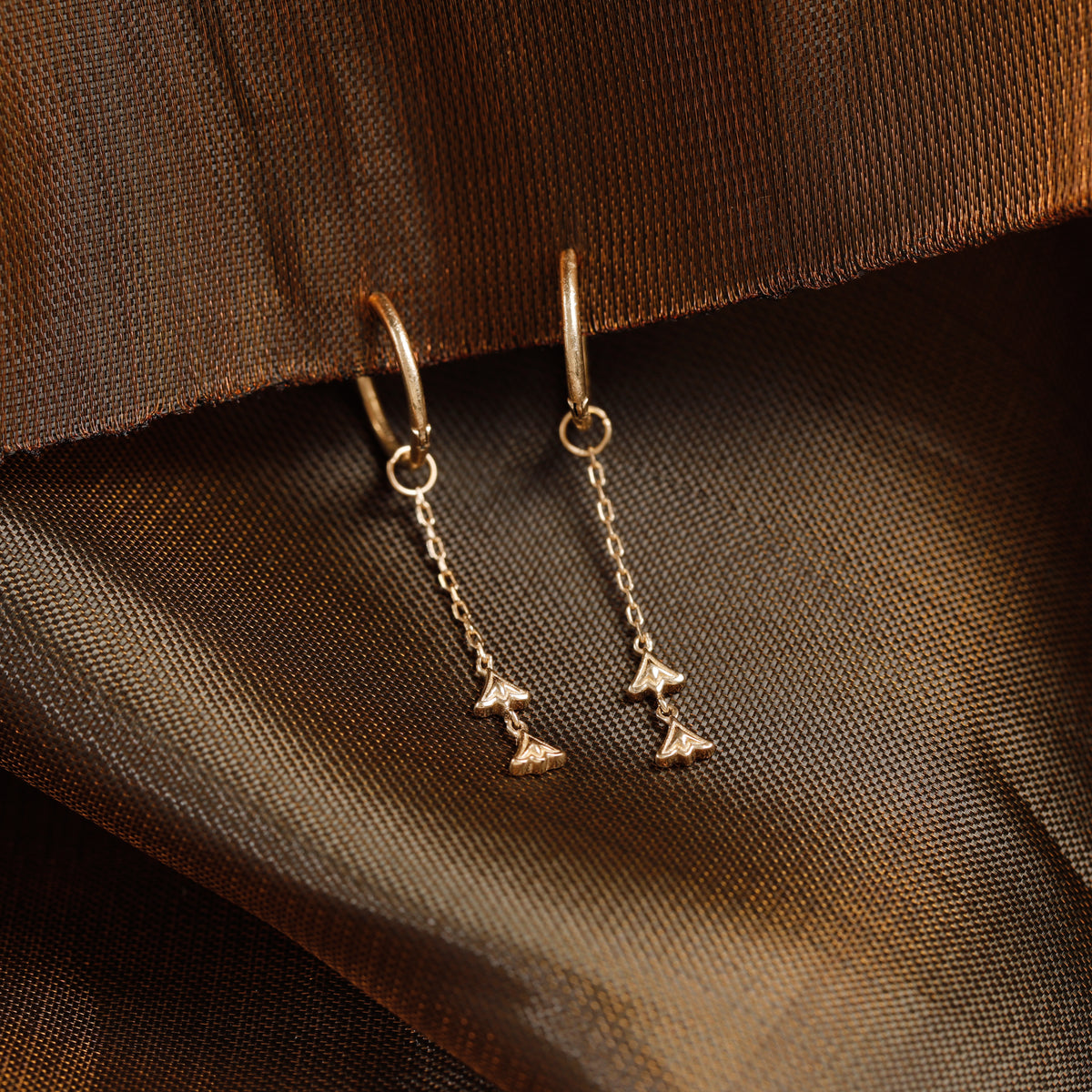 A pair of gold-toned dangle earrings with small chain drops and triangular charms, photographed on draped brown fabric.