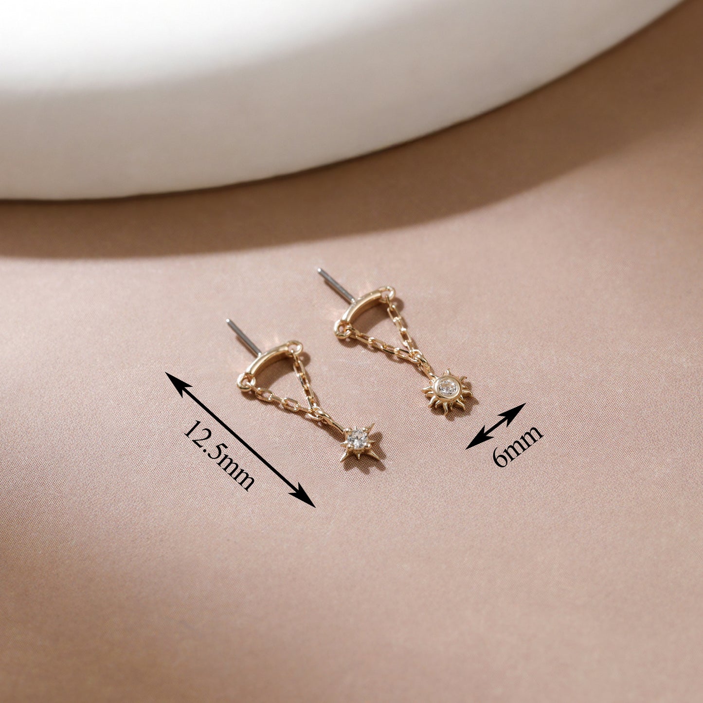 A pair of gold-toned hoop earrings with dangling star and sun motifs, shown on a beige surface with measurement arrows and labels indicating 12.5mm and 6mm dimensions.