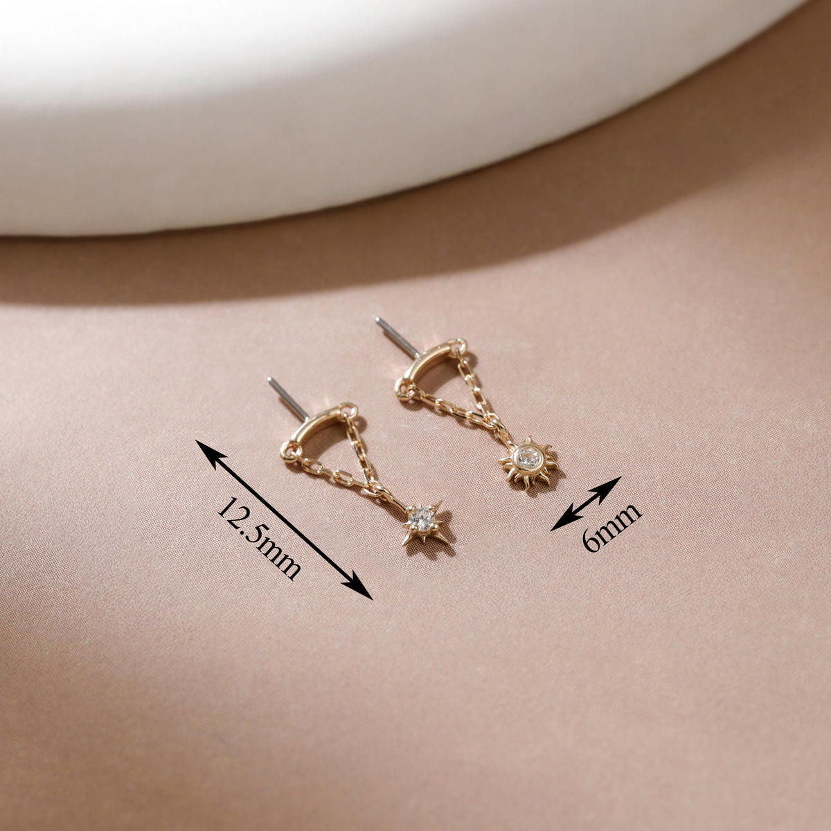 A pair of gold-toned hoop earrings with dangling star and sun motifs, shown on a beige surface with measurement arrows and labels indicating 12.5mm and 6mm dimensions.