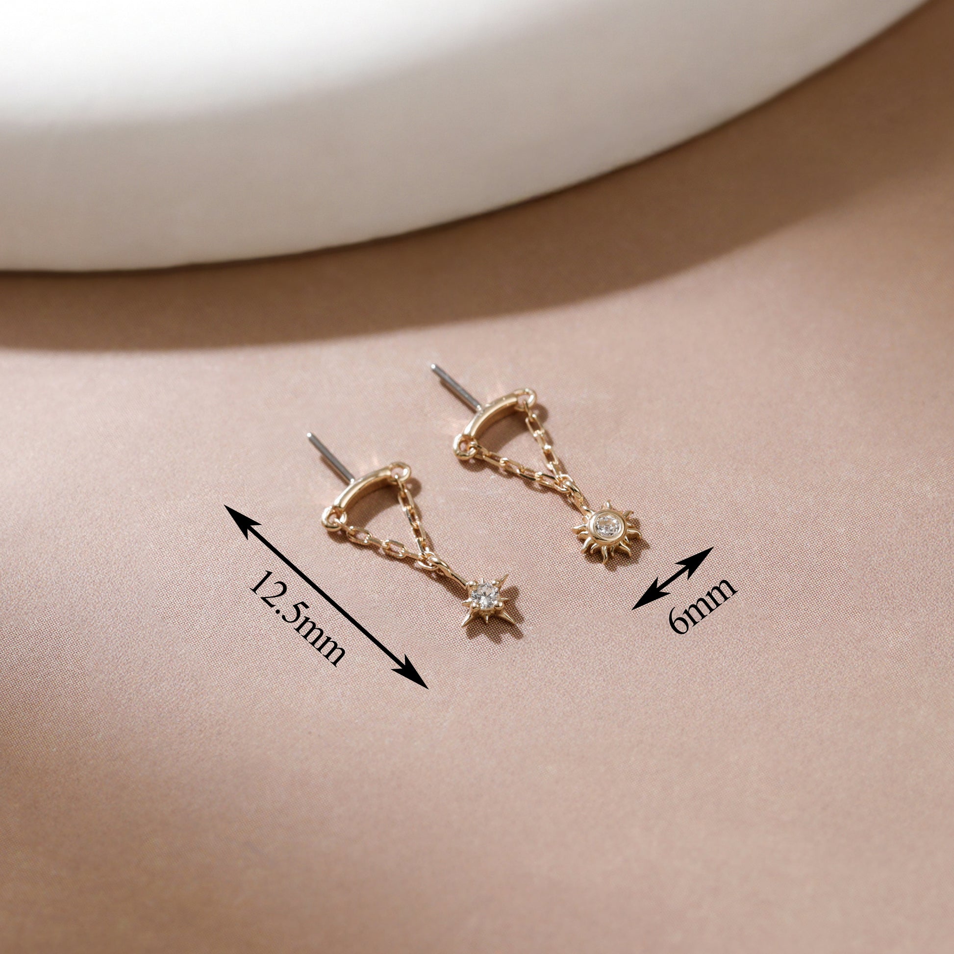 A pair of gold-toned hoop earrings with dangling star and sun motifs, shown on a beige surface with measurement arrows and labels indicating 12.5mm and 6mm dimensions.