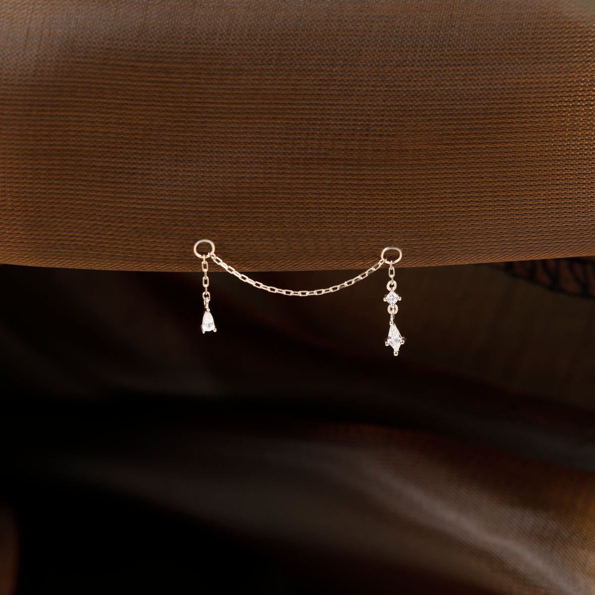 A delicate silver chain accessory with two dangling clear crystal teardrop charms, displayed against a textured brown fabric background.