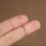 A close-up of a hand holding a delicate gold chain earring with a small dangling charm against a warm brown background.