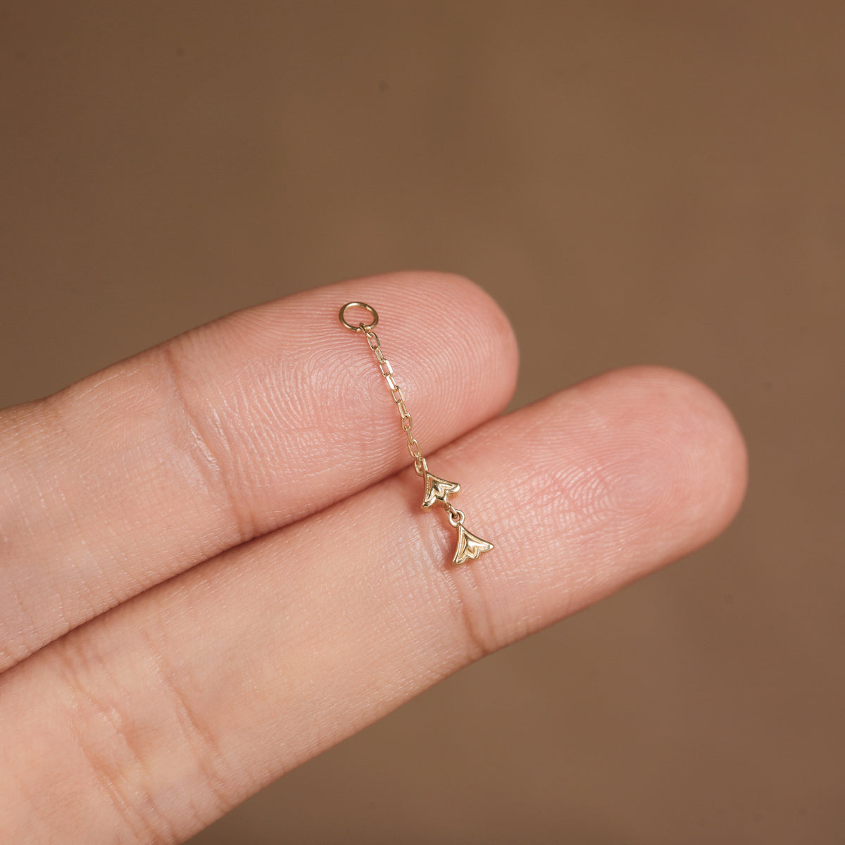 A close-up of a hand holding a delicate gold chain earring with a small dangling charm against a warm brown background.