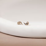 A pair of gold crescent moon stud earrings with delicate filigree detailing and small blue and clear gemstone accents, resting on a white curved surface.
