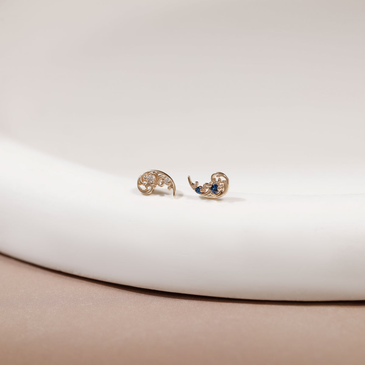 A pair of gold crescent moon stud earrings with delicate filigree detailing and small blue and clear gemstone accents, resting on a white curved surface.