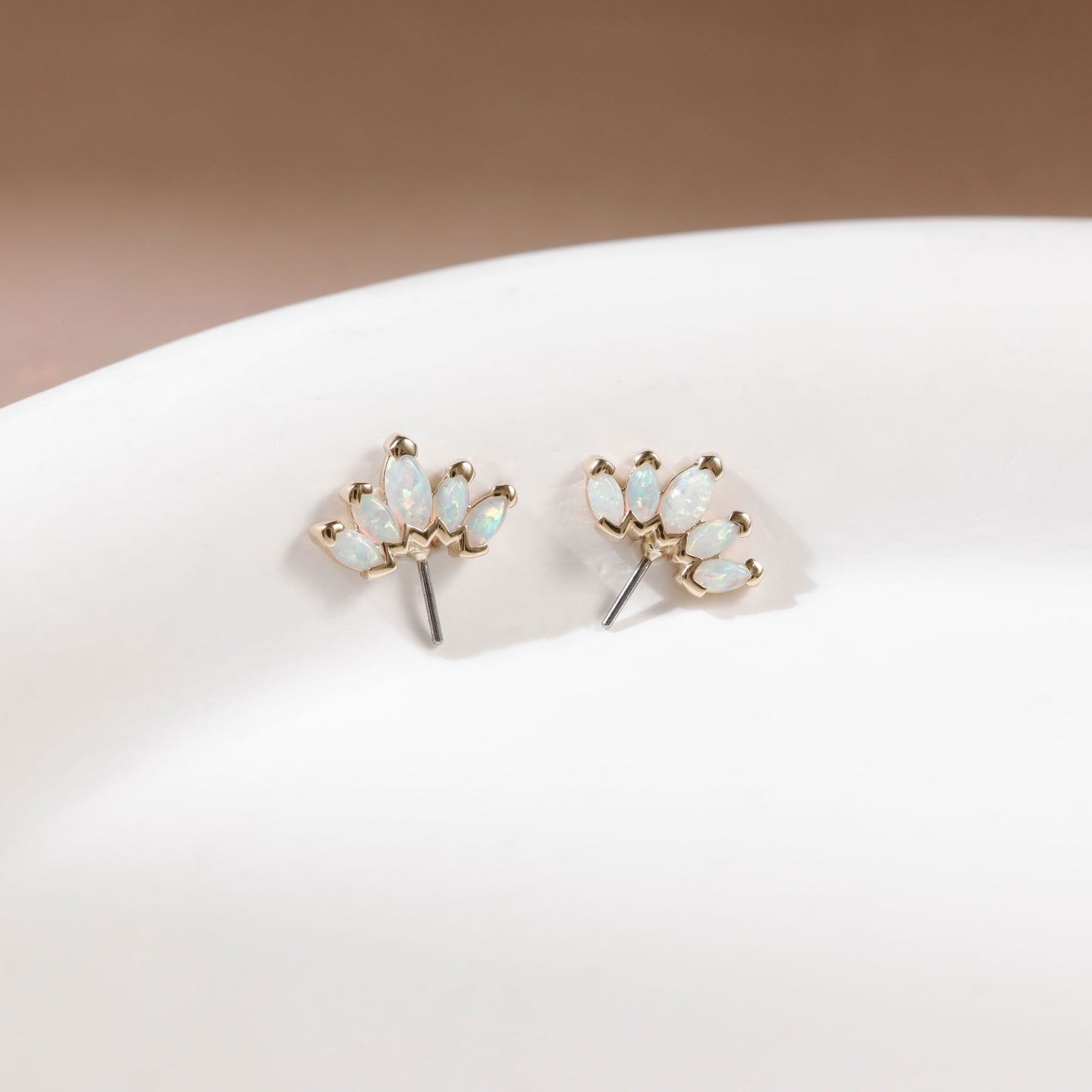 A pair of gold stud earrings with small iridescent white stones, displayed on a white curved surface against a soft beige background.