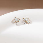 A pair of gold stud earrings with small iridescent white stones, displayed on a white curved surface against a soft beige background.