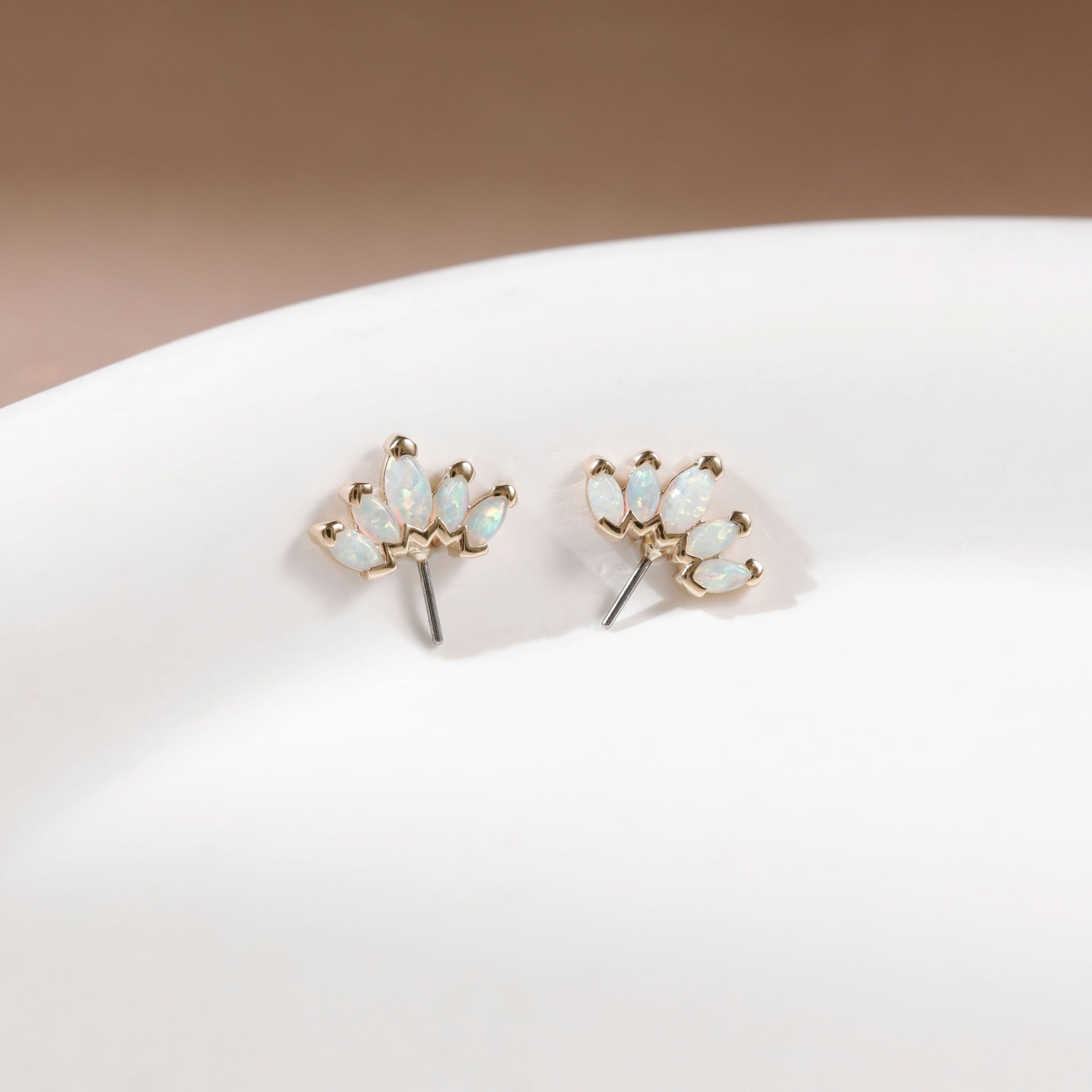 A pair of gold stud earrings with small iridescent white stones, displayed on a white curved surface against a soft beige background.