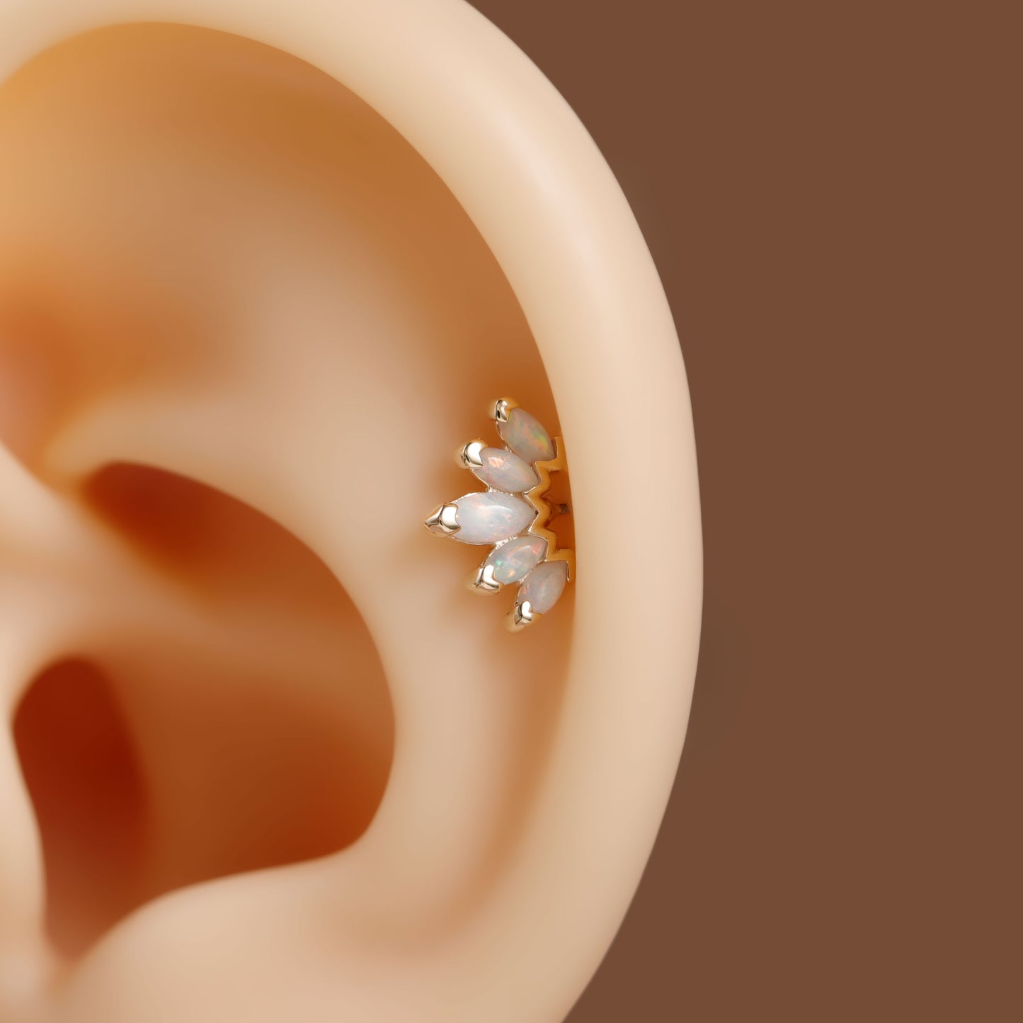 A close-up of a gold ear cuff with white pearl-like beads worn on a beige silicone ear model, set against a solid brown background.