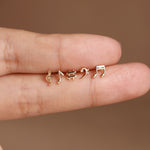 Four tiny gold-tone music note and symbol stud earrings displayed on a person's finger against a warm brown background.