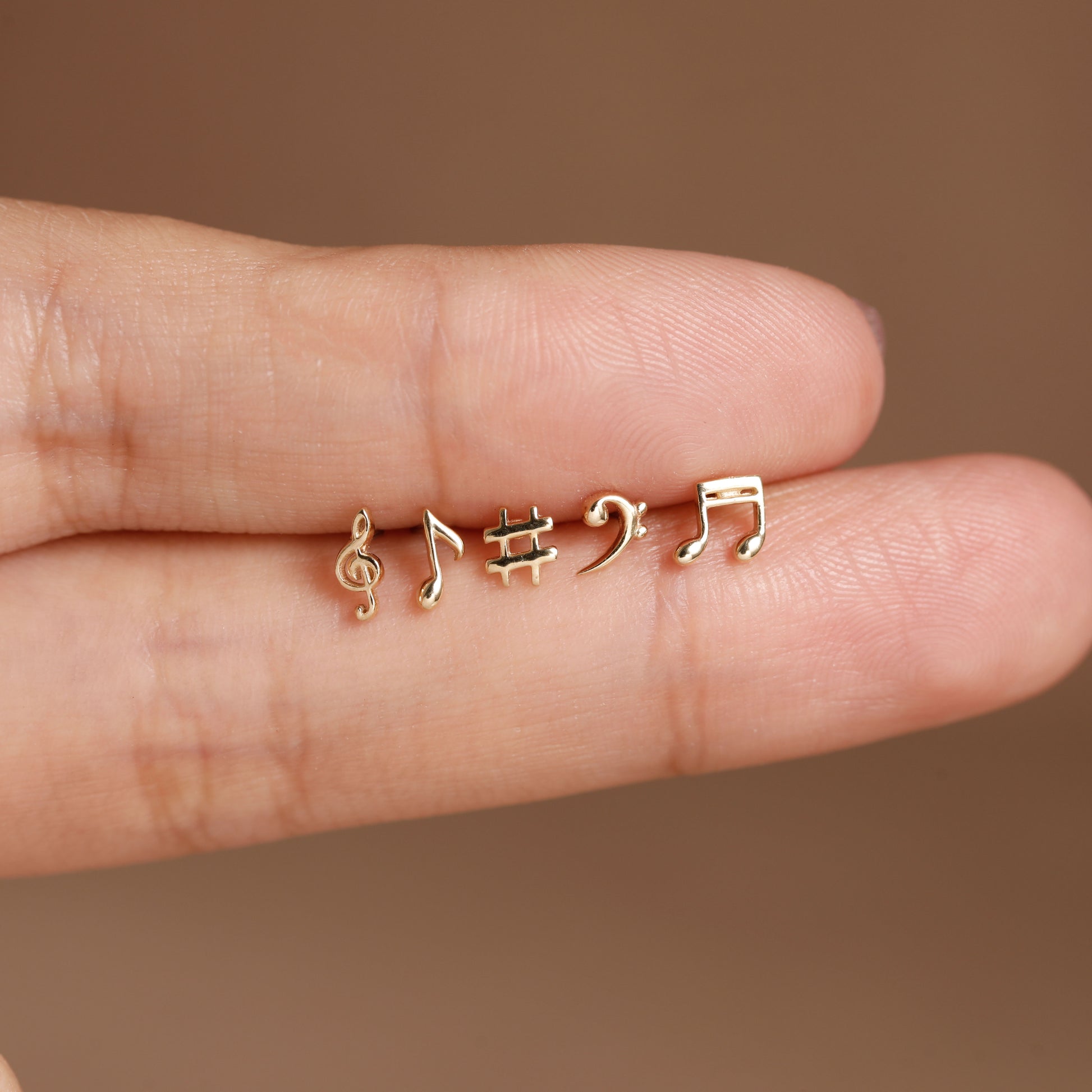 Four tiny gold-tone music note and symbol stud earrings displayed on a person's finger against a warm brown background.