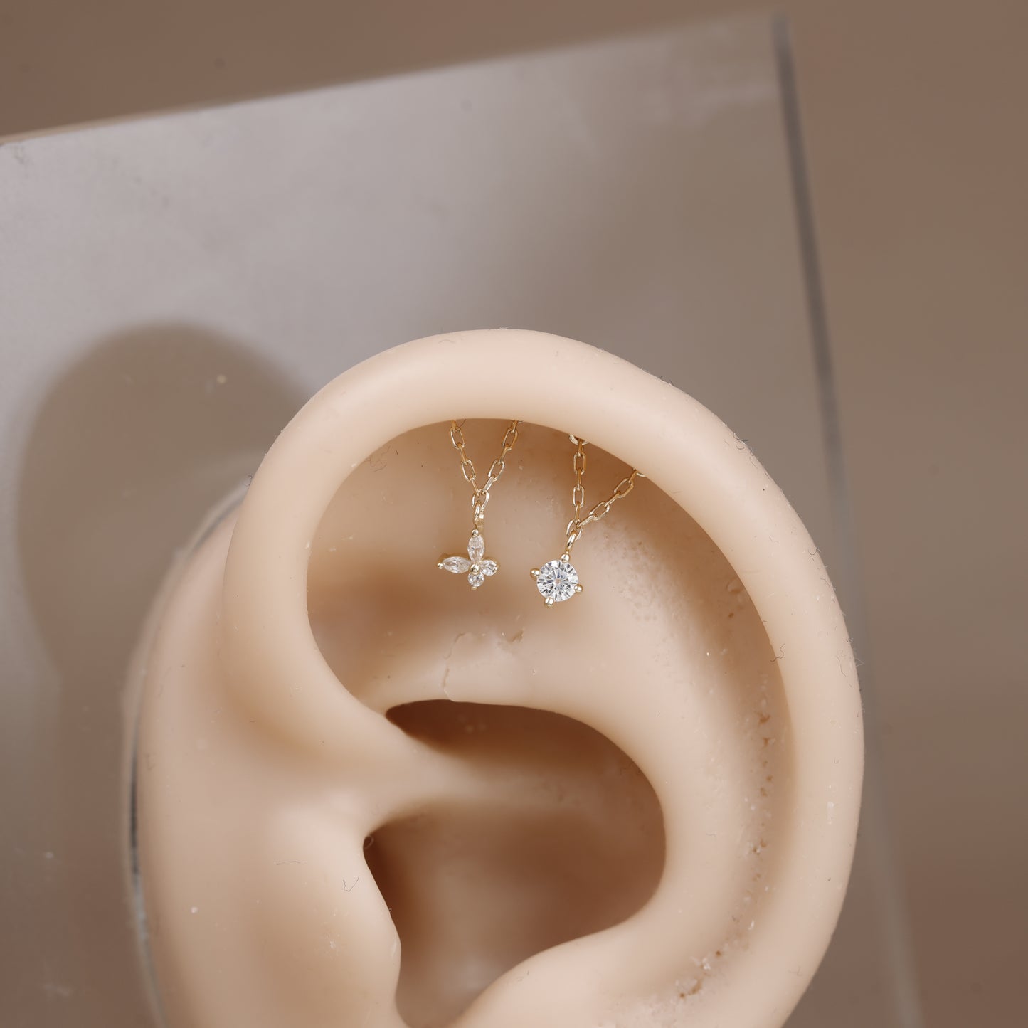 A pair of delicate gold-tone ear cuff earrings with clear gemstones displayed on a beige silicone ear model against a neutral beige background.
