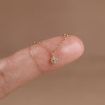 A delicate gold chain necklace with a small geometric starburst pendant set with clear stones, shown resting on a person's fingertip against a soft warm brown background.