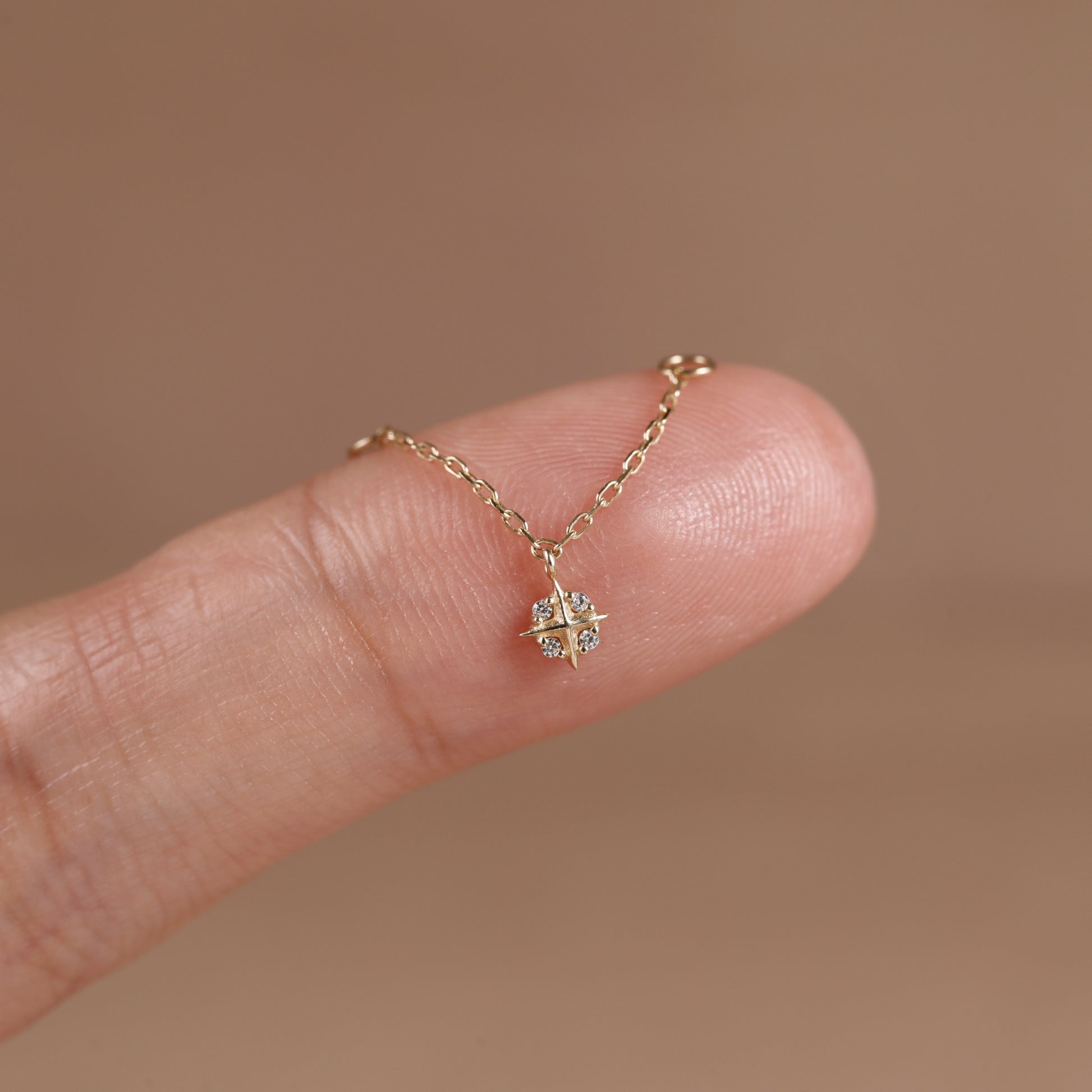 A delicate gold chain necklace with a small geometric starburst pendant set with clear stones, shown resting on a person's fingertip against a soft warm brown background.