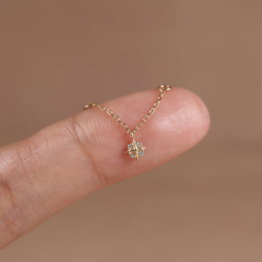A delicate gold chain necklace with a small geometric starburst pendant set with clear stones, shown resting on a person's fingertip against a soft warm brown background.