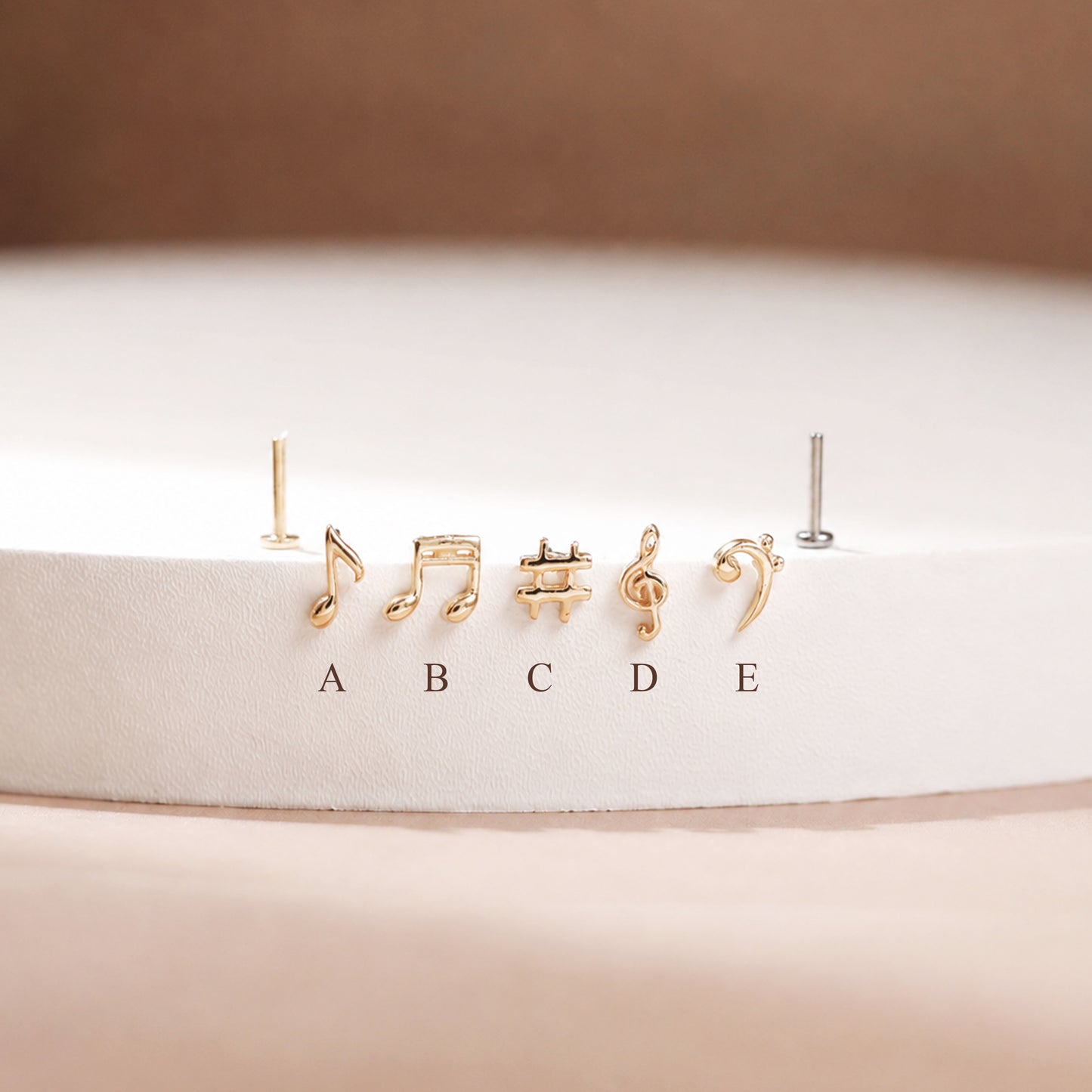 Five small gold-tone stud earrings shaped like musical notes and symbols, displayed on a white circular pedestal with labels A through E below them. One earring on the left is shown without a post, and one on the right appears silver-toned.