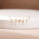 Five small gold-tone stud earrings shaped like musical notes and symbols, displayed on a white circular pedestal with labels A through E below them. One earring on the left is shown without a post, and one on the right appears silver-toned.