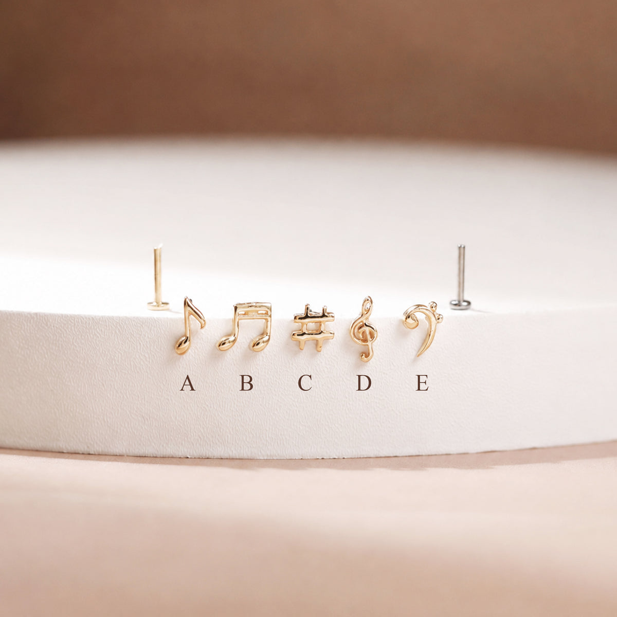 Five small gold-tone stud earrings shaped like musical notes and symbols, displayed on a white circular pedestal with labels A through E below them. One earring on the left is shown without a post, and one on the right appears silver-toned.