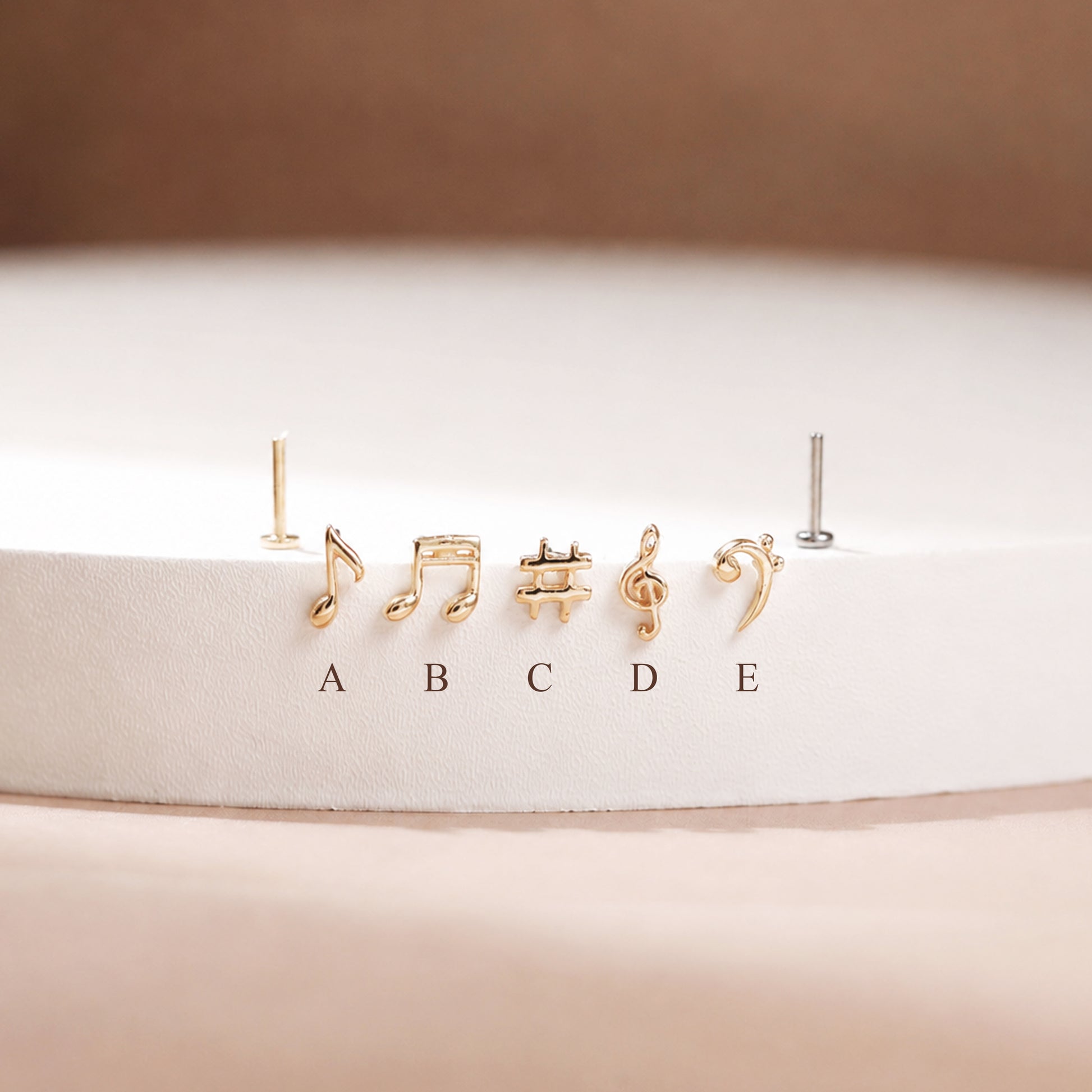 Five small gold-tone stud earrings shaped like musical notes and symbols, displayed on a white circular pedestal with labels A through E below them. One earring on the left is shown without a post, and one on the right appears silver-toned.