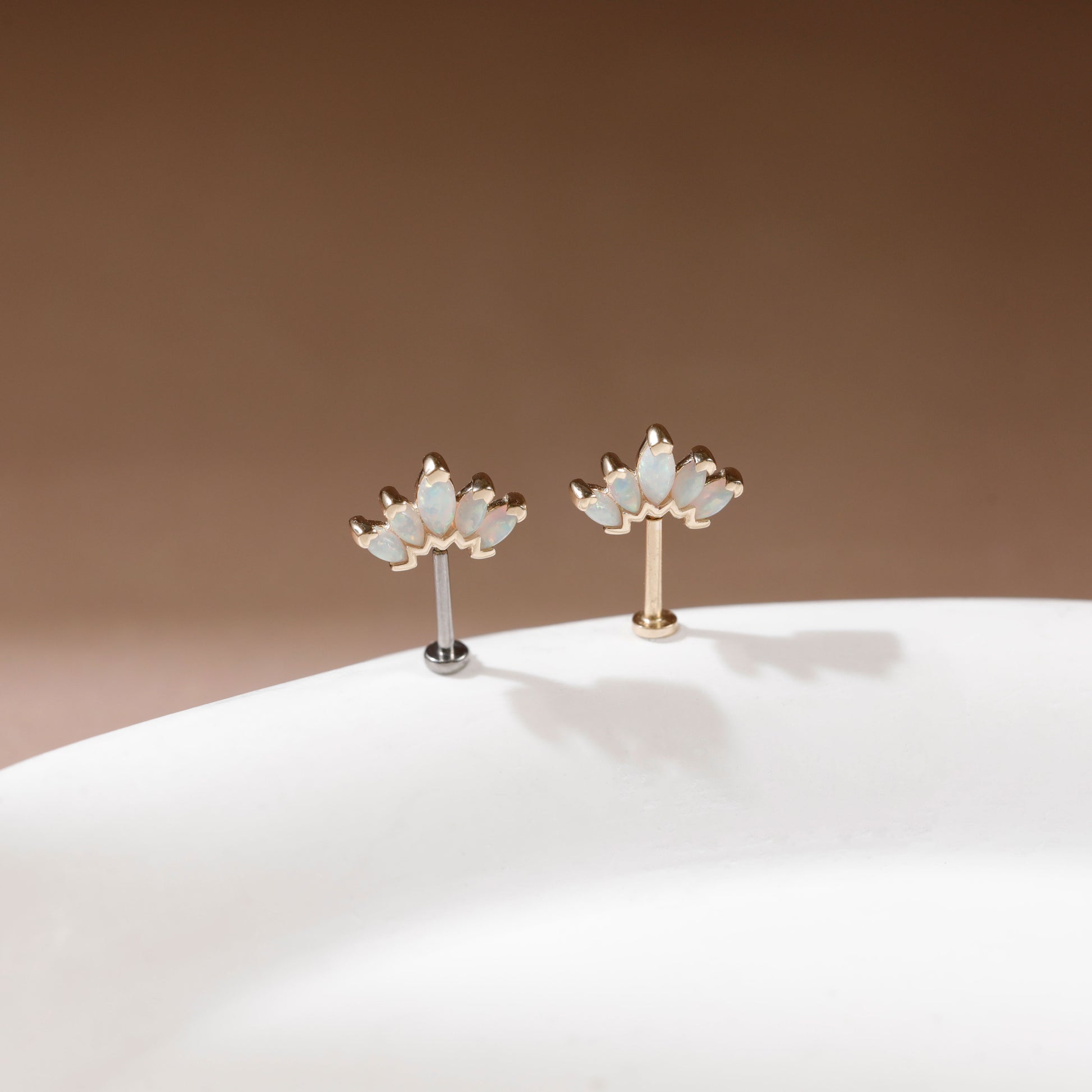 A pair of gold and white lotus flower stud earrings displayed on a white curved surface against a warm brown background.