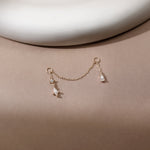 A delicate gold chain accessory with two small teardrop-shaped clear crystal pendants, displayed on a warm beige surface with a white curved background element.