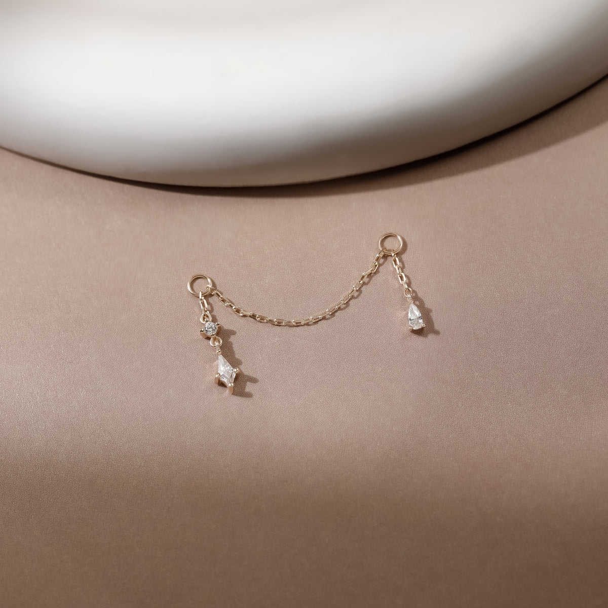 A delicate gold chain accessory with two small teardrop-shaped clear crystal pendants, displayed on a warm beige surface with a white curved background element.
