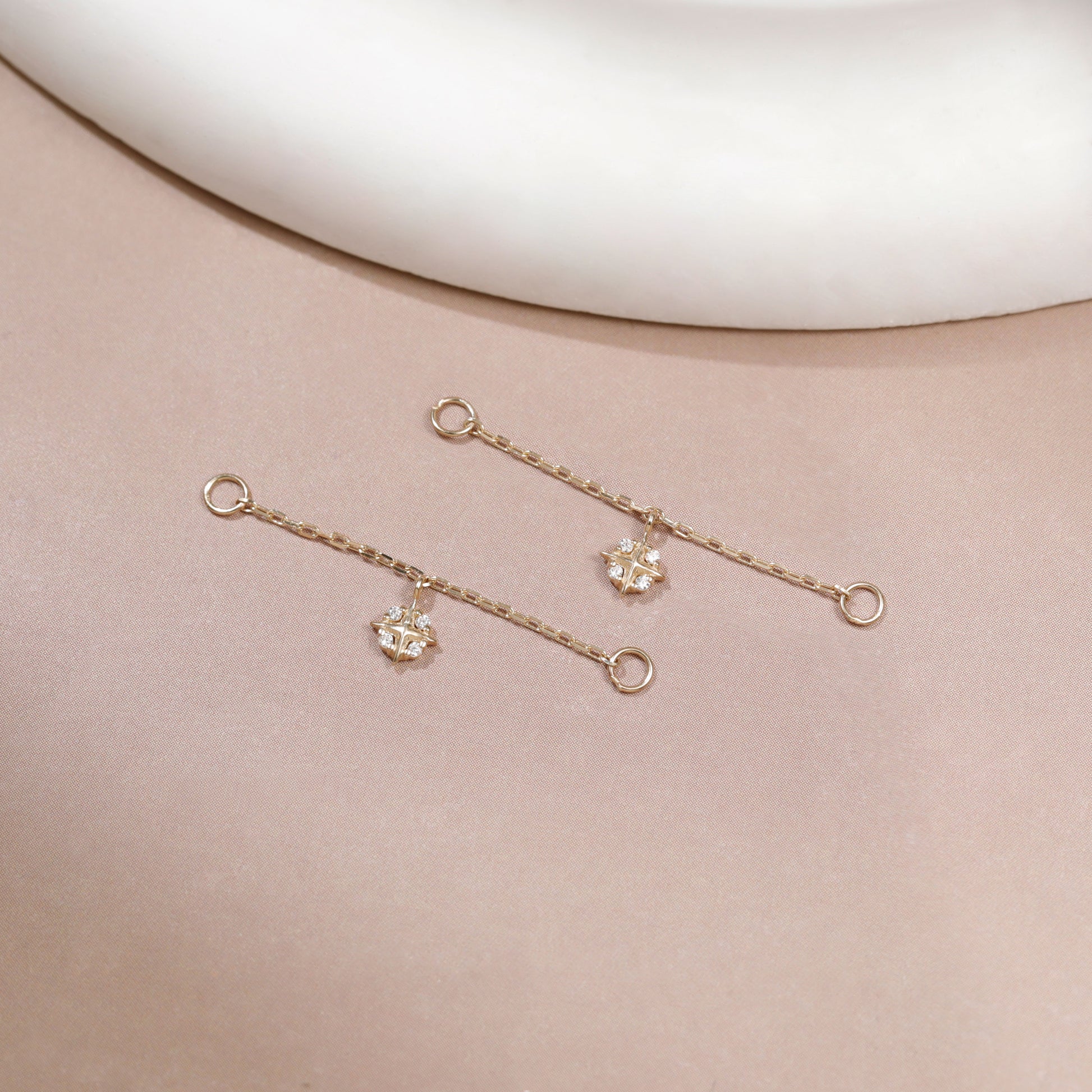A pair of delicate gold dangle earrings with small clear crystal charms, displayed on a soft beige surface with a white curved prop at the top.