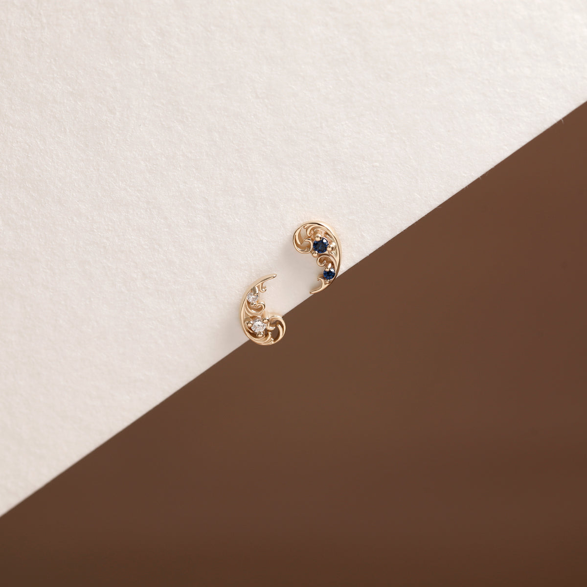 A pair of gold crescent-shaped hoop earrings with small dark blue gemstones and clear accent stones, displayed on a diagonally split cream and brown surface.