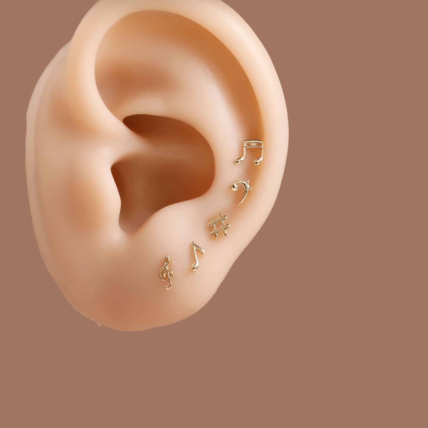A close-up of a human ear wearing several small gold ear piercings, including music note and curved stud designs, against a solid warm brown background.