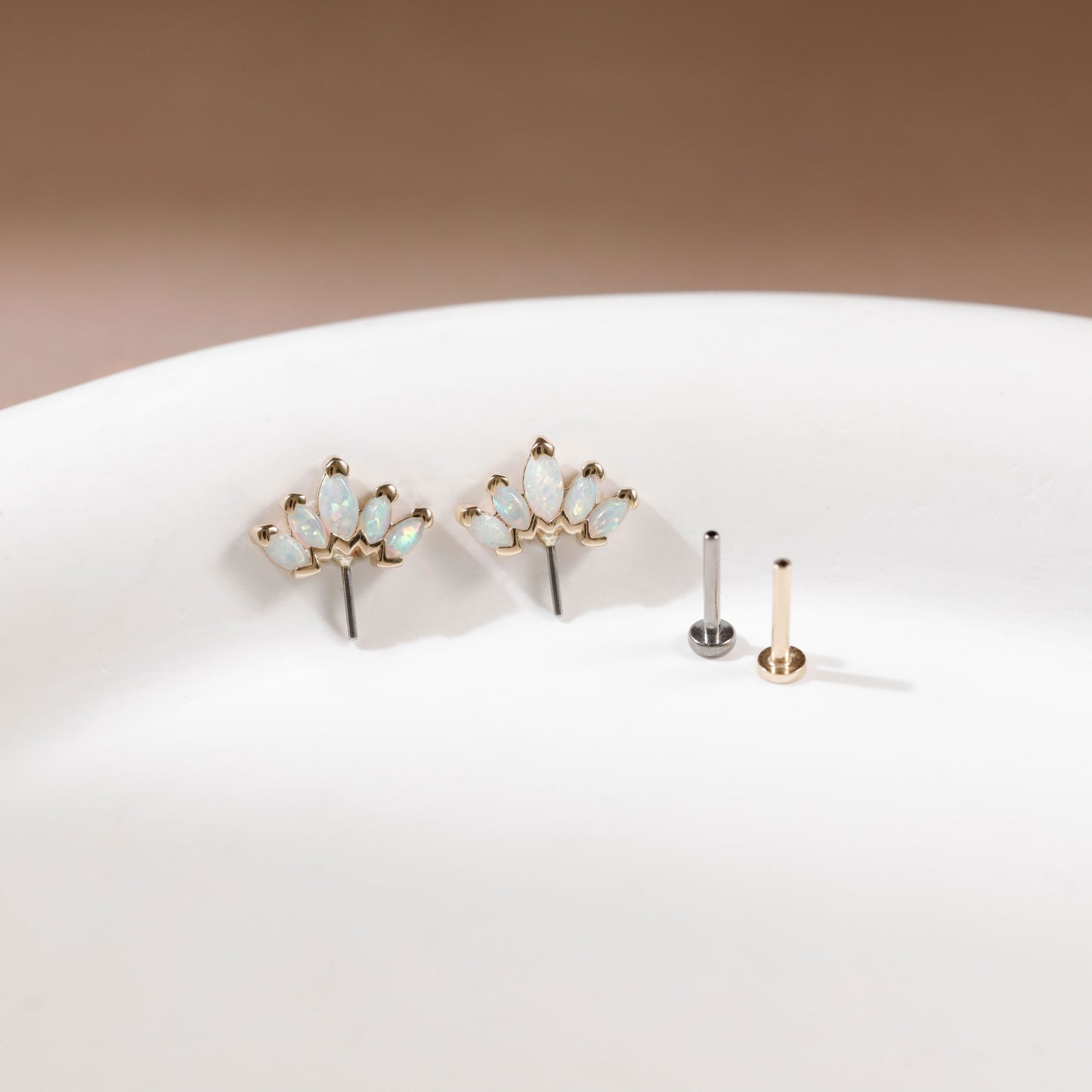 Gold floral stud earrings with small iridescent opal-like stones, displayed on a white surface next to two flat backings, one silver and one gold, against a warm brown background.