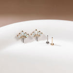 Gold floral stud earrings with small iridescent opal-like stones, displayed on a white surface next to two flat backings, one silver and one gold, against a warm brown background.