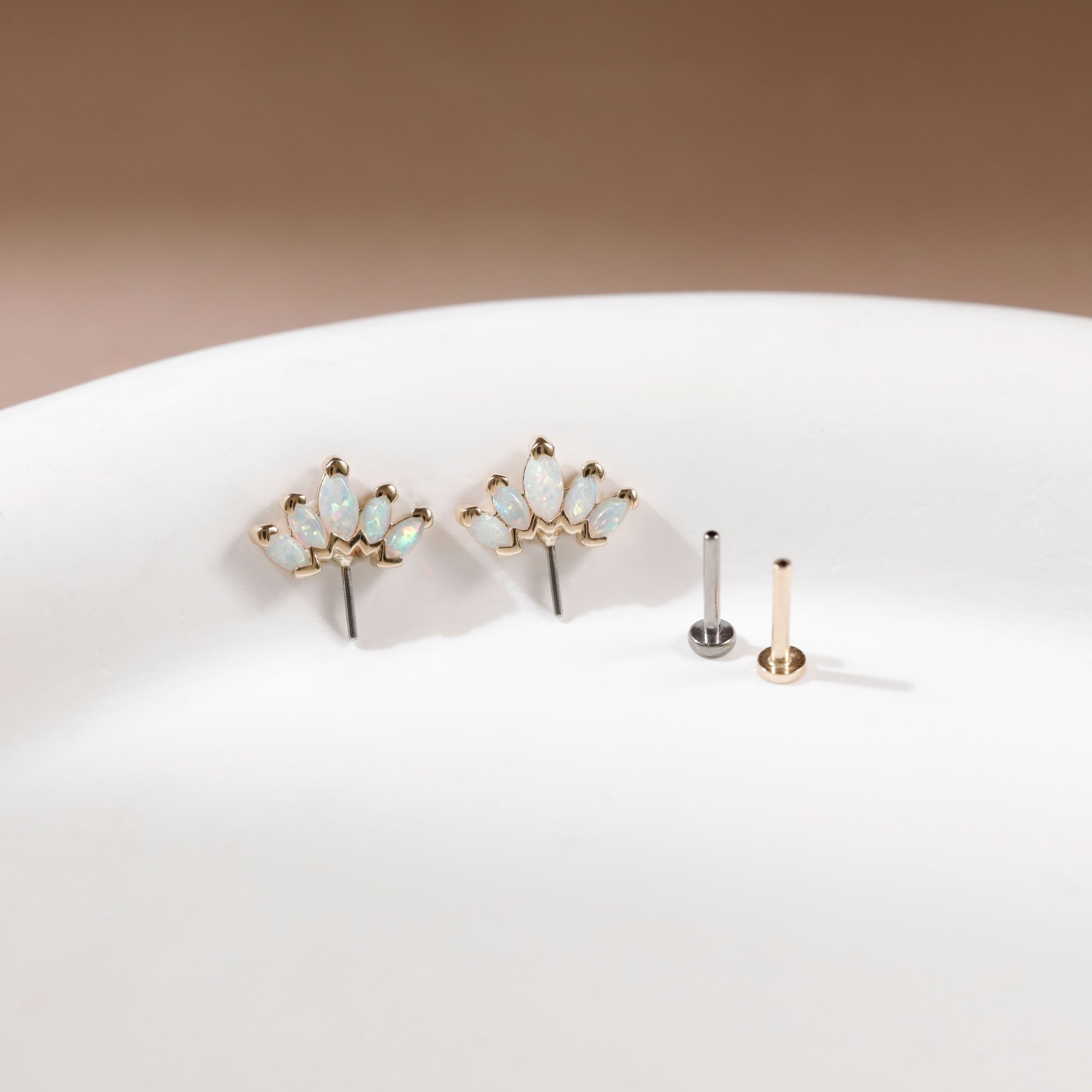 Gold floral stud earrings with small iridescent opal-like stones, displayed on a white surface next to two flat backings, one silver and one gold, against a warm brown background.