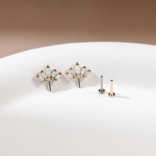 Gold floral stud earrings with small iridescent opal-like stones, displayed on a white surface next to two flat backings, one silver and one gold, against a warm brown background.