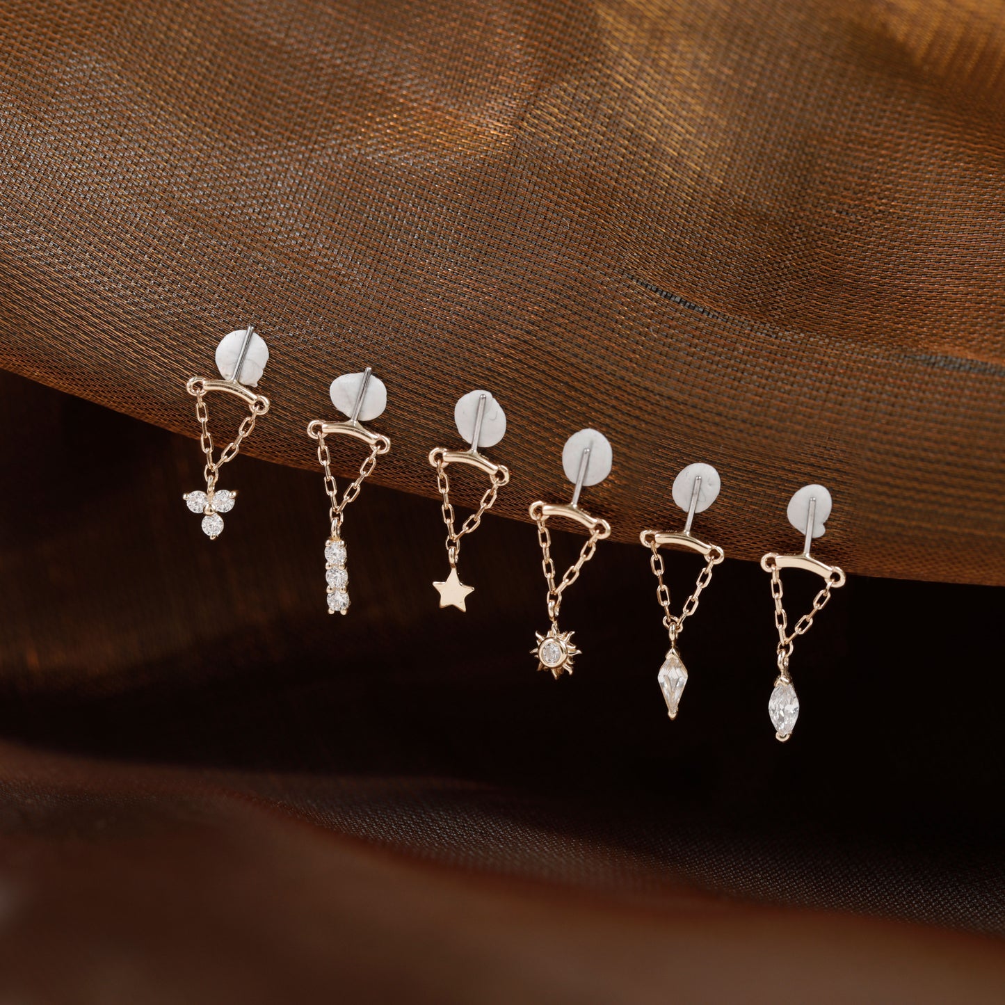 Six small gold-toned dangle earrings with clear stones and celestial motifs are arranged on a draped brown fabric background.