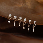 Six small gold-toned dangle earrings with clear stones and celestial motifs are arranged on a draped brown fabric background.