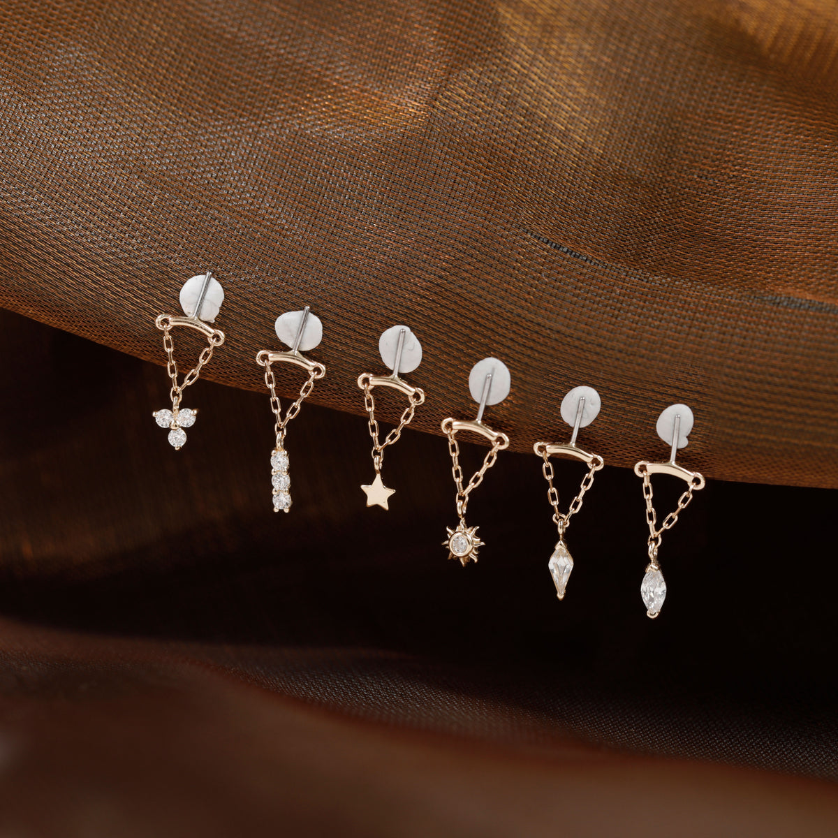 Six small gold-toned dangle earrings with clear stones and celestial motifs are arranged on a draped brown fabric background.