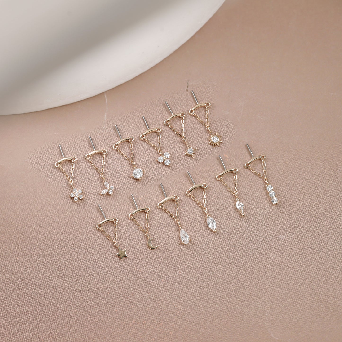 Twelve gold-toned ear piercings with various celestial and floral charms, arranged on a soft beige surface with a white curved prop in the background.