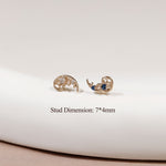 A pair of gold crescent-shaped stud earrings with ornate scrollwork and small blue gemstone accents, displayed on a white curved surface with text reading 'Stud Dimension: 7*4mm'.