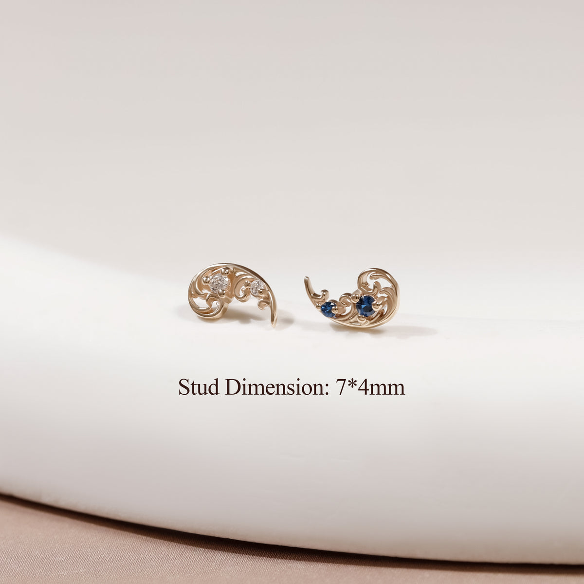 A pair of gold crescent-shaped stud earrings with ornate scrollwork and small blue gemstone accents, displayed on a white curved surface with text reading 'Stud Dimension: 7*4mm'.