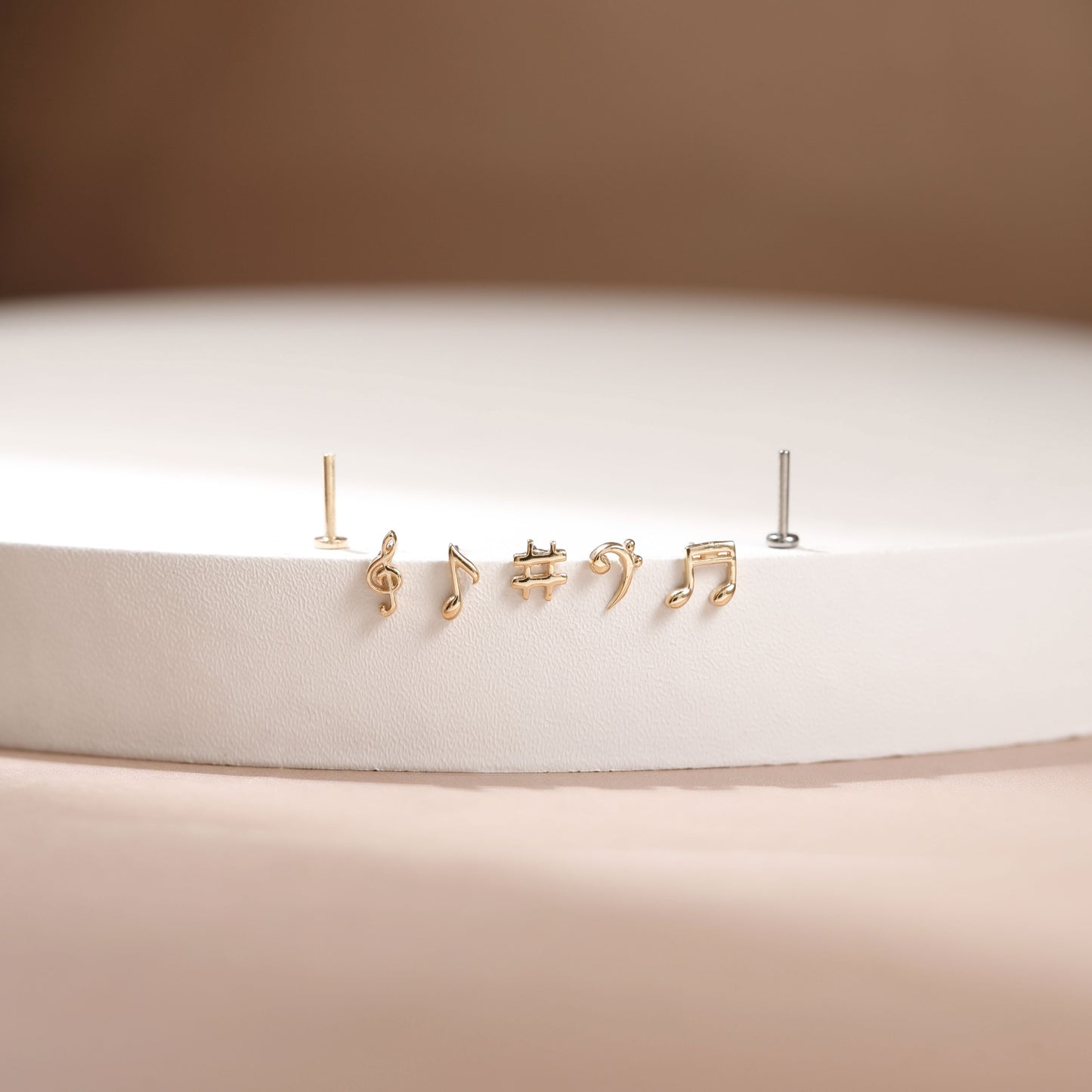 A pair of gold stud earrings shaped like musical notes, displayed on a white circular pedestal against a warm beige background.