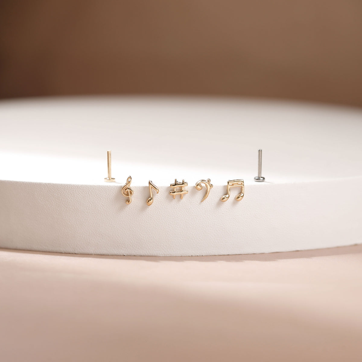 A pair of gold stud earrings shaped like musical notes, displayed on a white circular pedestal against a warm beige background.