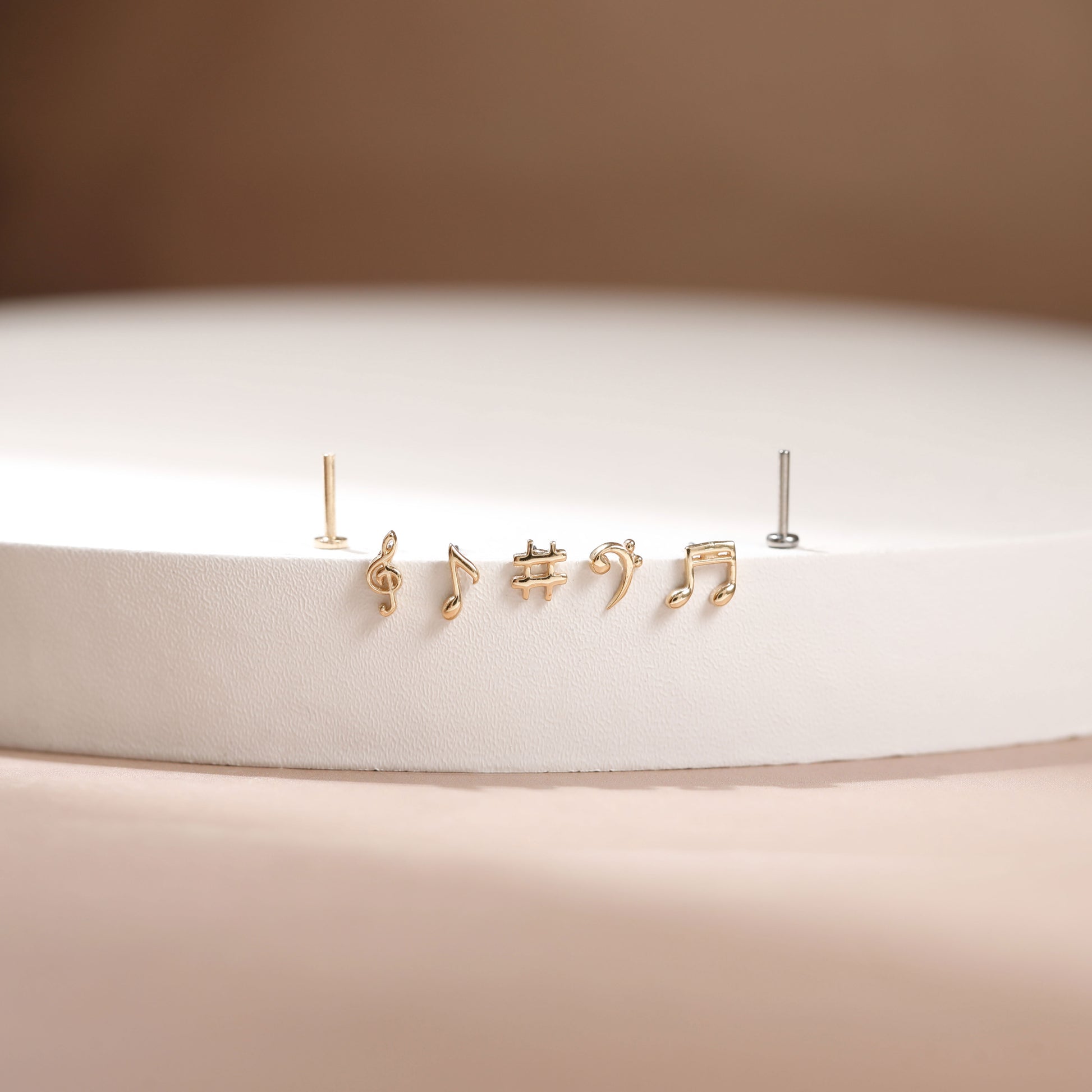 A pair of gold stud earrings shaped like musical notes, displayed on a white circular pedestal against a warm beige background.