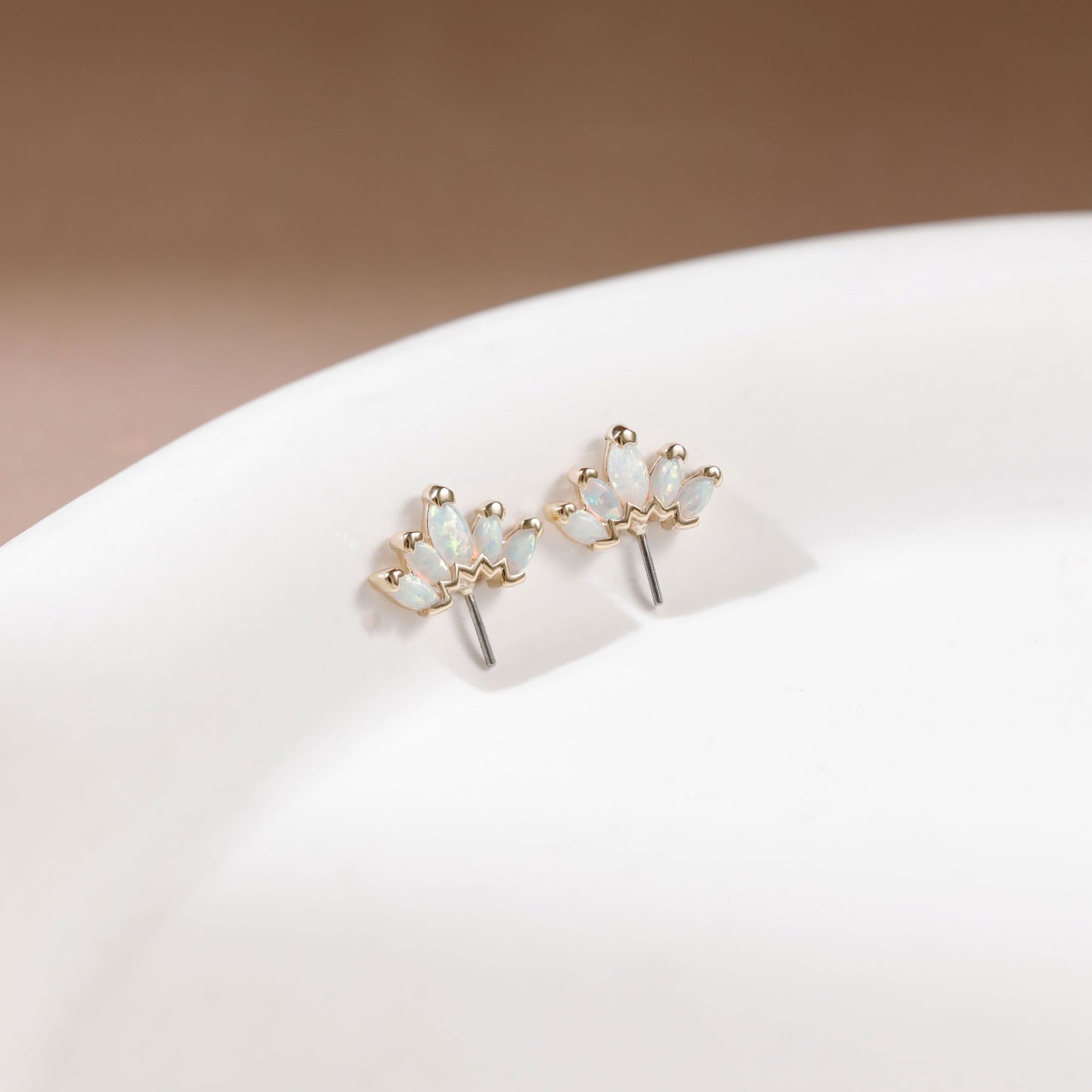 A pair of gold flower-shaped stud earrings with iridescent white opal-like stones, displayed on a white ceramic surface against a soft beige background.