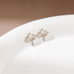 A pair of gold flower-shaped stud earrings with iridescent white opal-like stones, displayed on a white ceramic surface against a soft beige background.