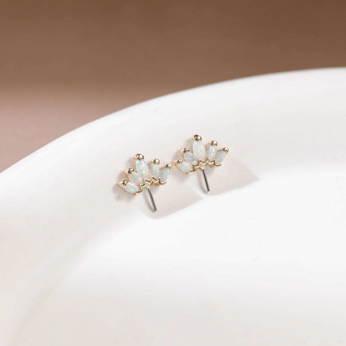 A pair of gold flower-shaped stud earrings with iridescent white opal-like stones, displayed on a white ceramic surface against a soft beige background.