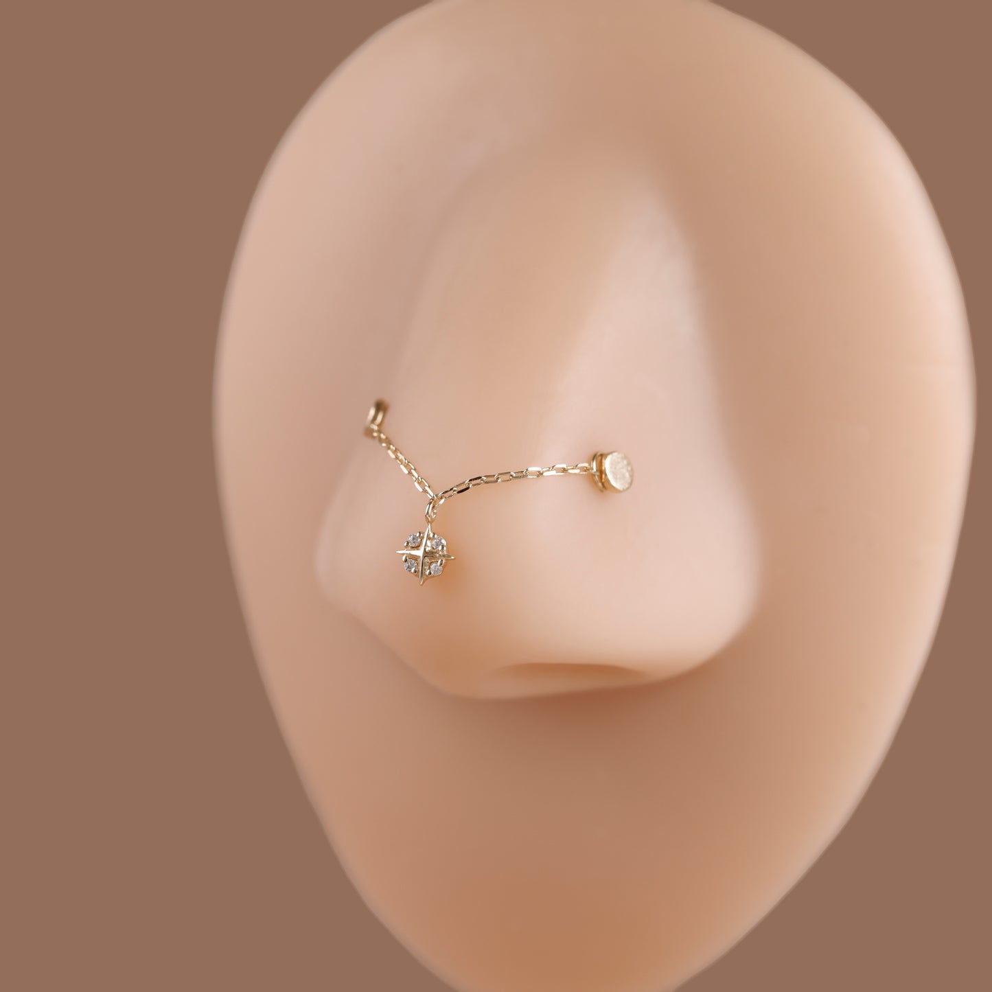 A delicate gold nose stud with a small dangling crystal cluster and a tiny round accent, displayed on a beige silicone nose model against a solid taupe background.