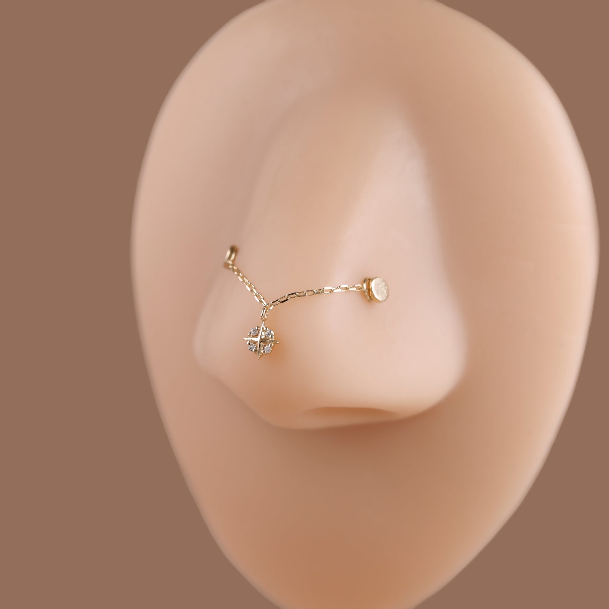 A delicate gold nose stud with a small dangling crystal cluster and a tiny round accent, displayed on a beige silicone nose model against a solid taupe background.